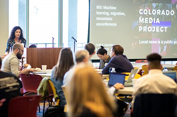 A woman presenting at a podium in front of a crowd of people with a slideshow with the Colorado Media Project logo being projected