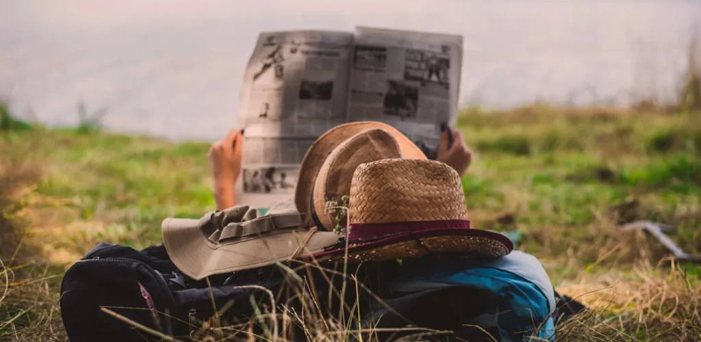 A woman laying on her backpack in a grassy field while reading a newspaper