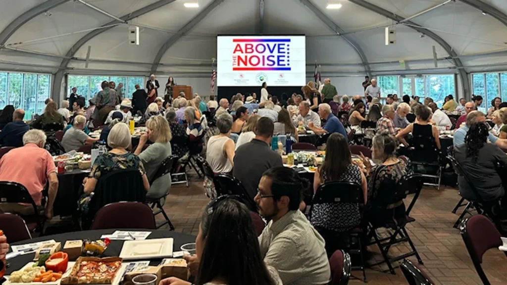 A large crowd sitting at tables while a screen projects the "Above the Noise" logo
