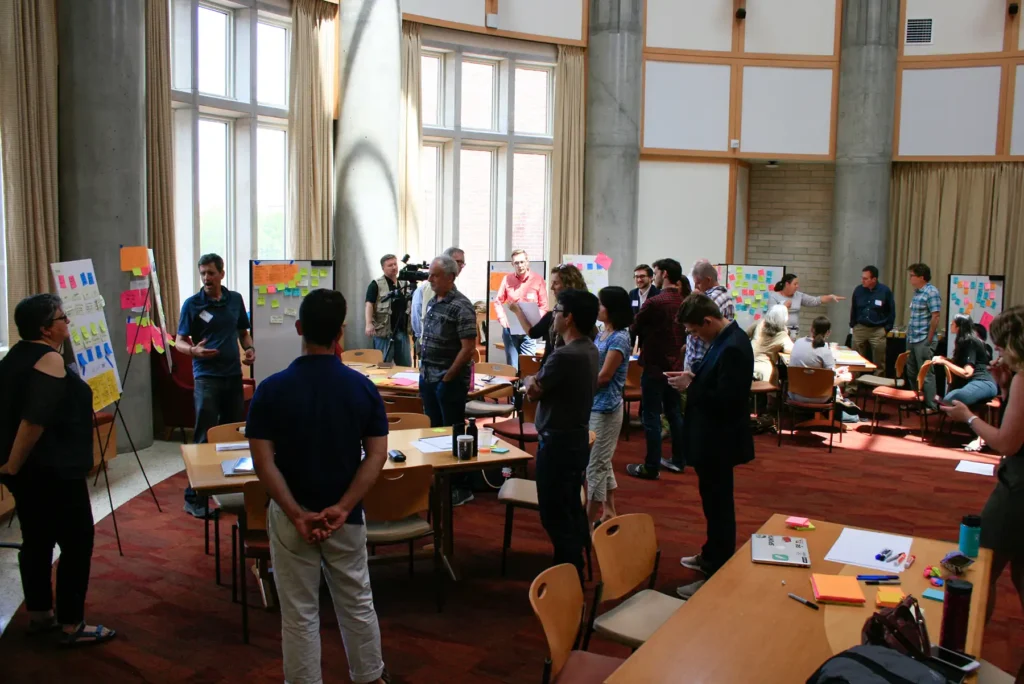 A group of people gather around multiple whiteboards spread throughout a room with colorful sticky notes on them