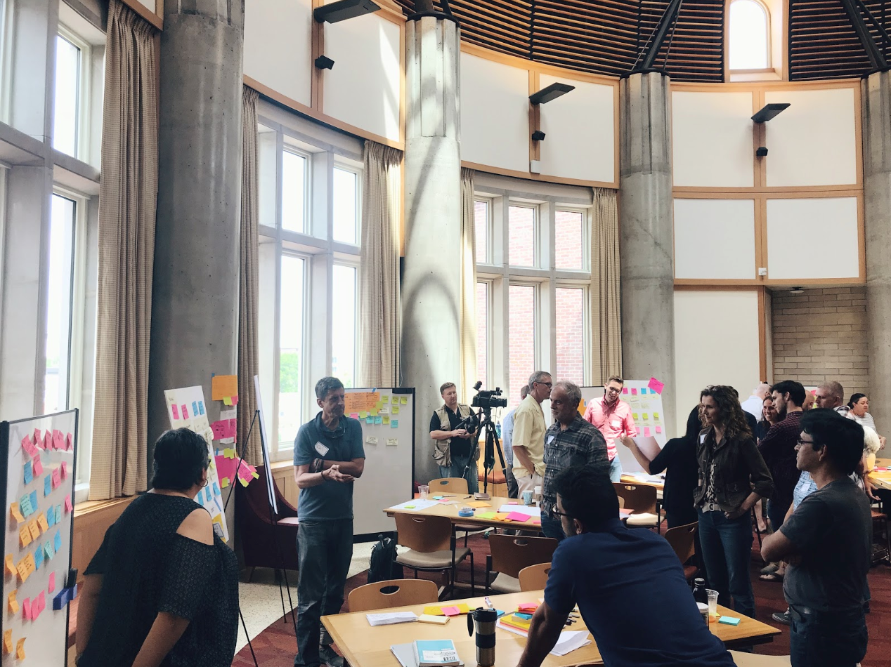 A group of people gather in a large room with whiteboards and sticky notes of various colors on them
