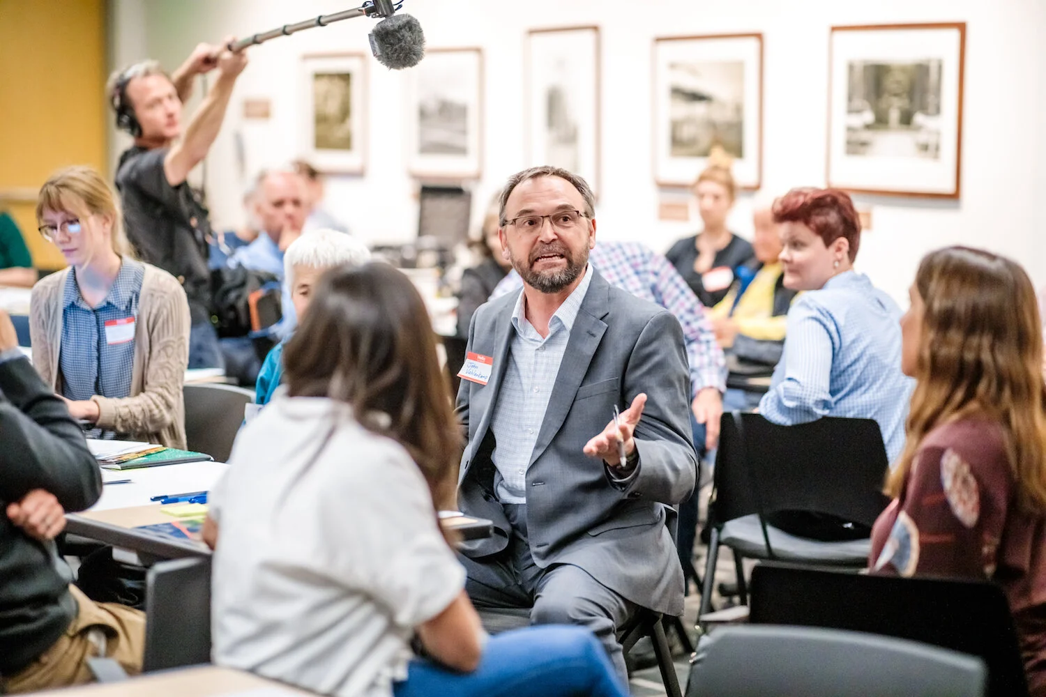 A man speaks to a group of people while another man holding a boom microphone