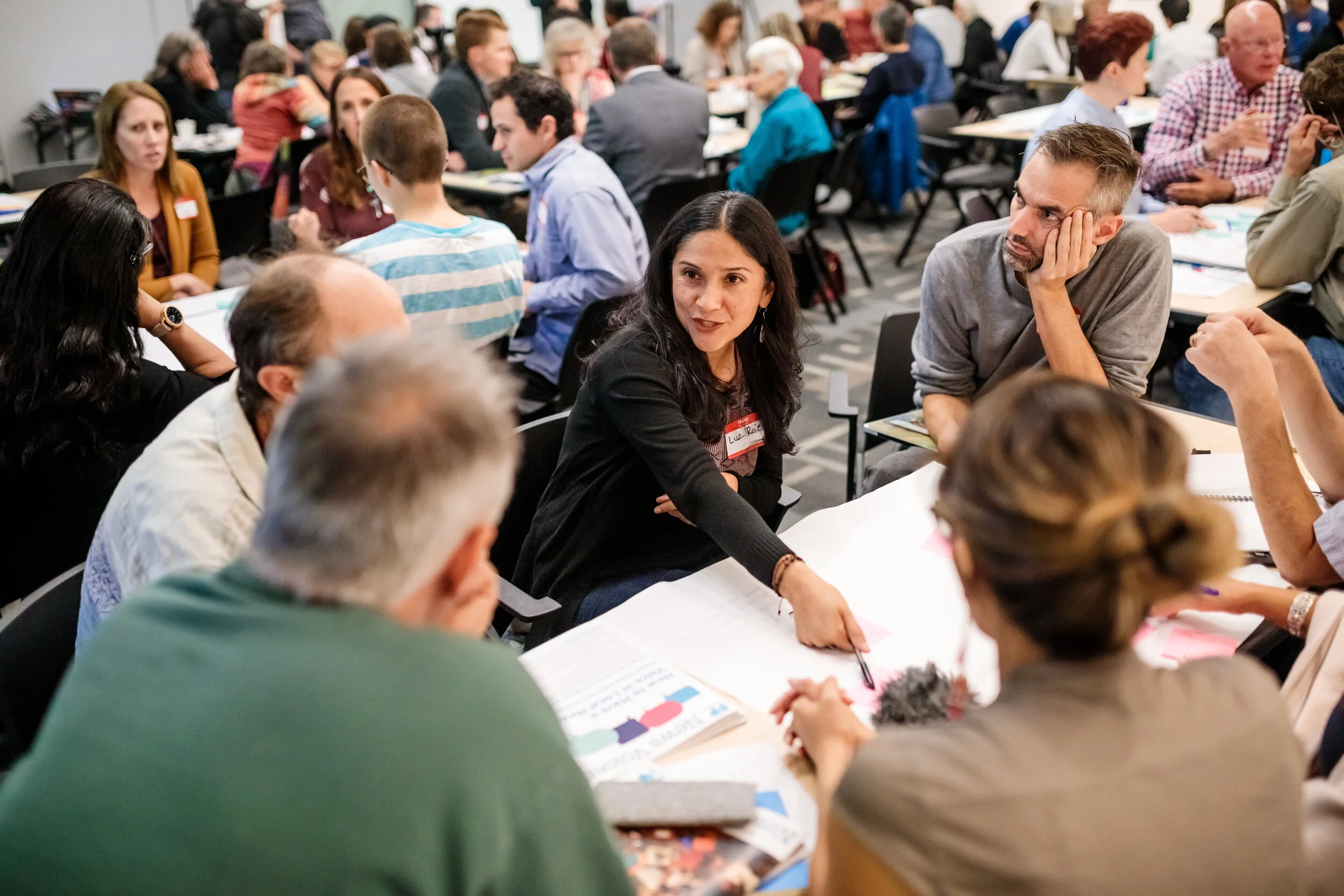 A woman points at a sticky note of a table while surrounded by a group of other people in a busy room.