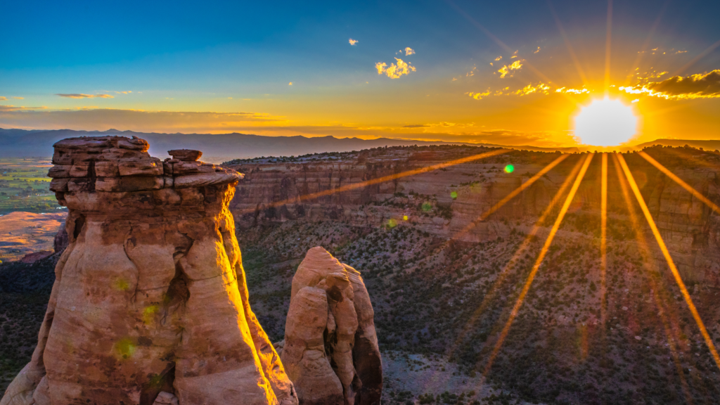 The sun sets over a rugged landscape of canyons and rock formations, casting long rays of light across the scene.